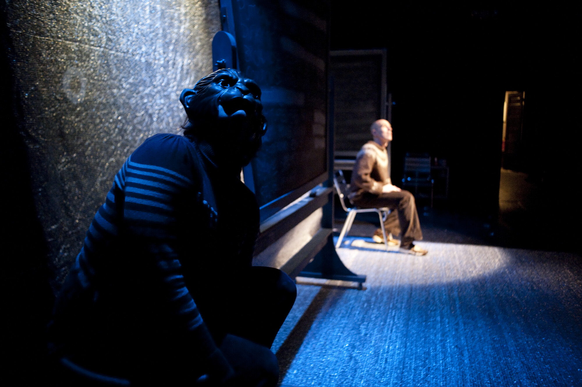 A performer in a monkey mask sits offstage while another performer sits under a spotlight on stage.