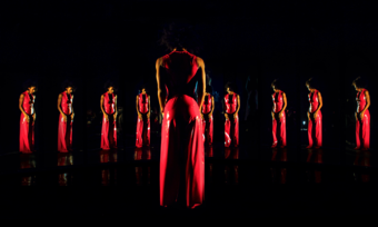 performer in a red dress looking at multiple reflections with herself in a mirror 