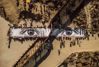 An aerial photograph of a picnic table spanning the US Mexico border wall with people eating on either side.