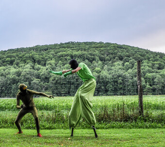 Outside in a field, performer leans away from another stilt-wearing performer.