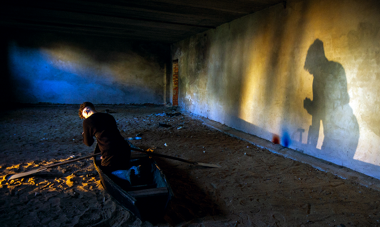 a person crouching in a boat in a large, empty room with concrete walls and dirt floor. their shadow is on the wall to their right