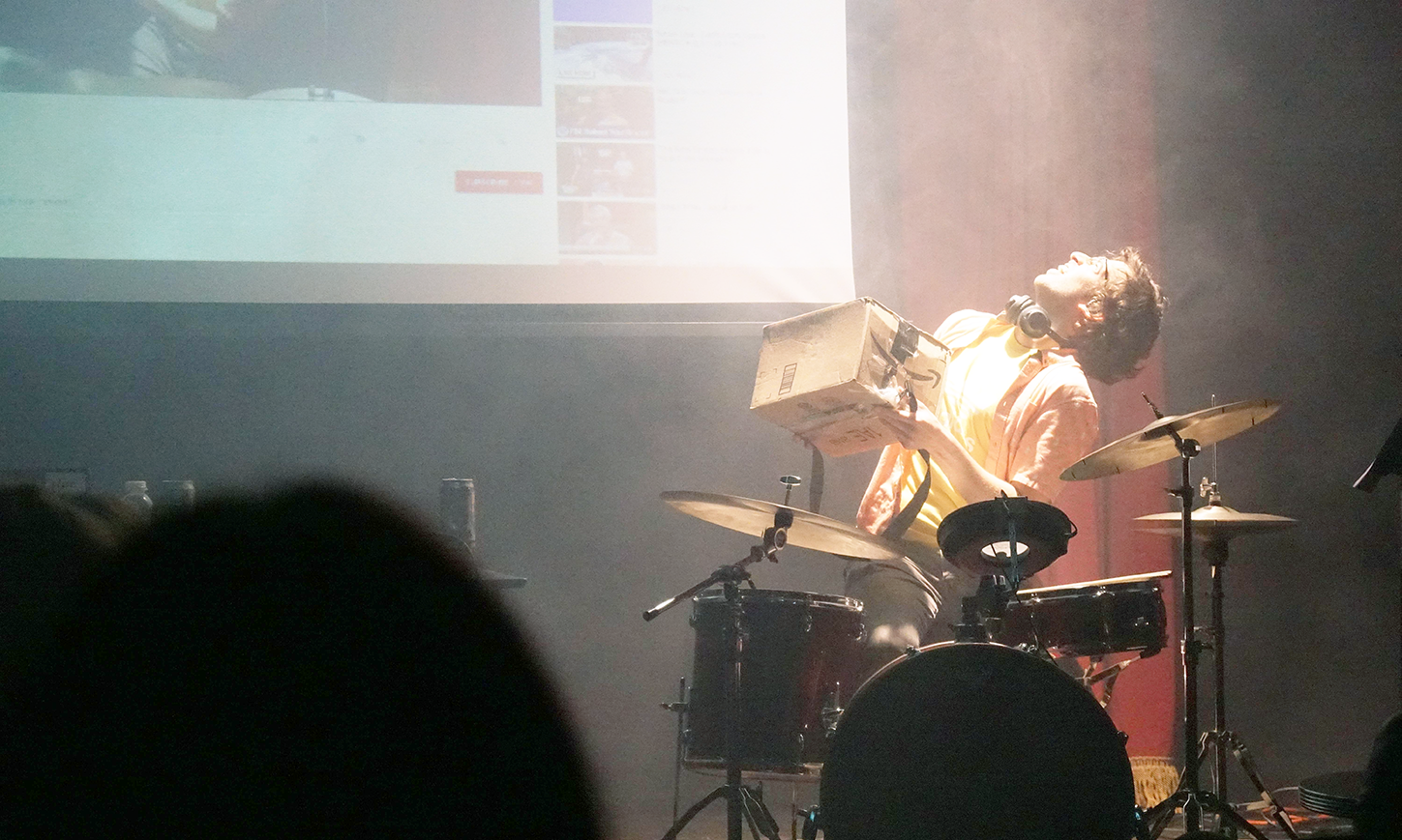 a person seated onstage at a drumset looking up at a projection