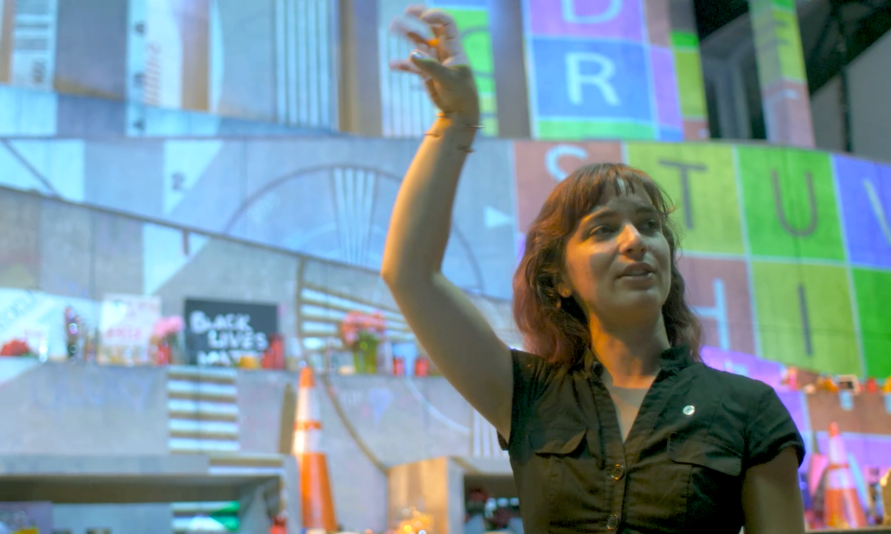 Katherine Freer, a white woman with long brown hair wearing a black dress, gestures at the slightly out of focus stage for a production of Antigone, where the architecture is illuminated by the projections of a colorful focus grid.