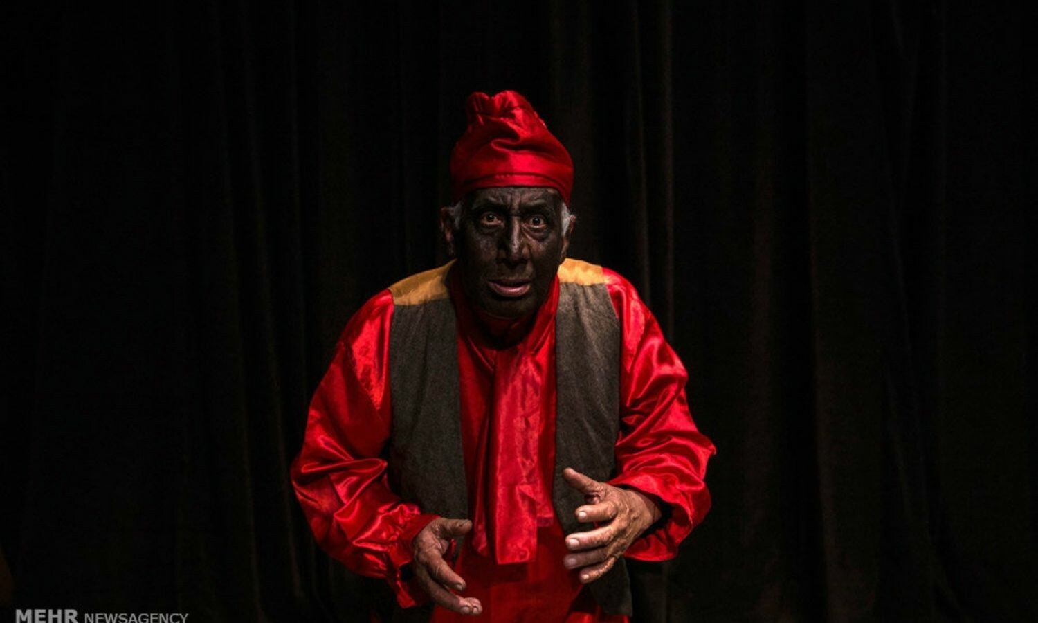 An Image of a man standing on stage slight hunched wearing black face, satin red cap, a read blouse, and brown vest. 