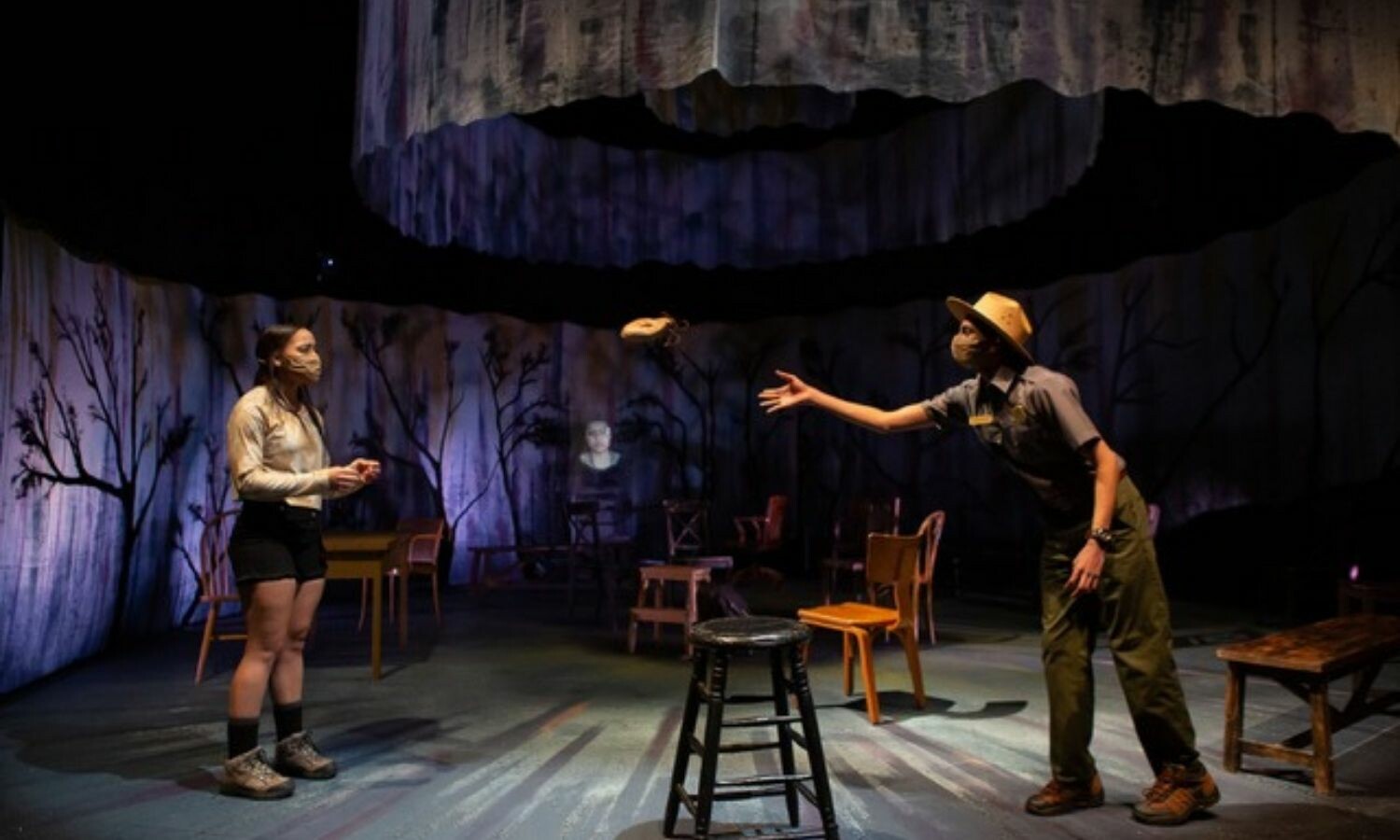 A man reaching out to a woman from whom he's distanced. Both are surrounded by chairs and the onstage "woods".