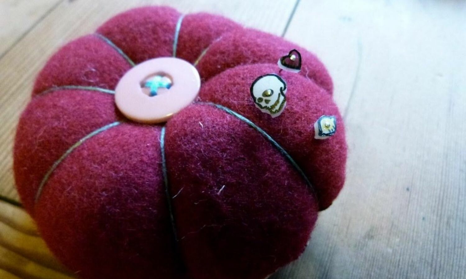 Close up image of a red pin cushion. Pinned to the cushion are small plastic images of a heart, a skull, and a lemon.