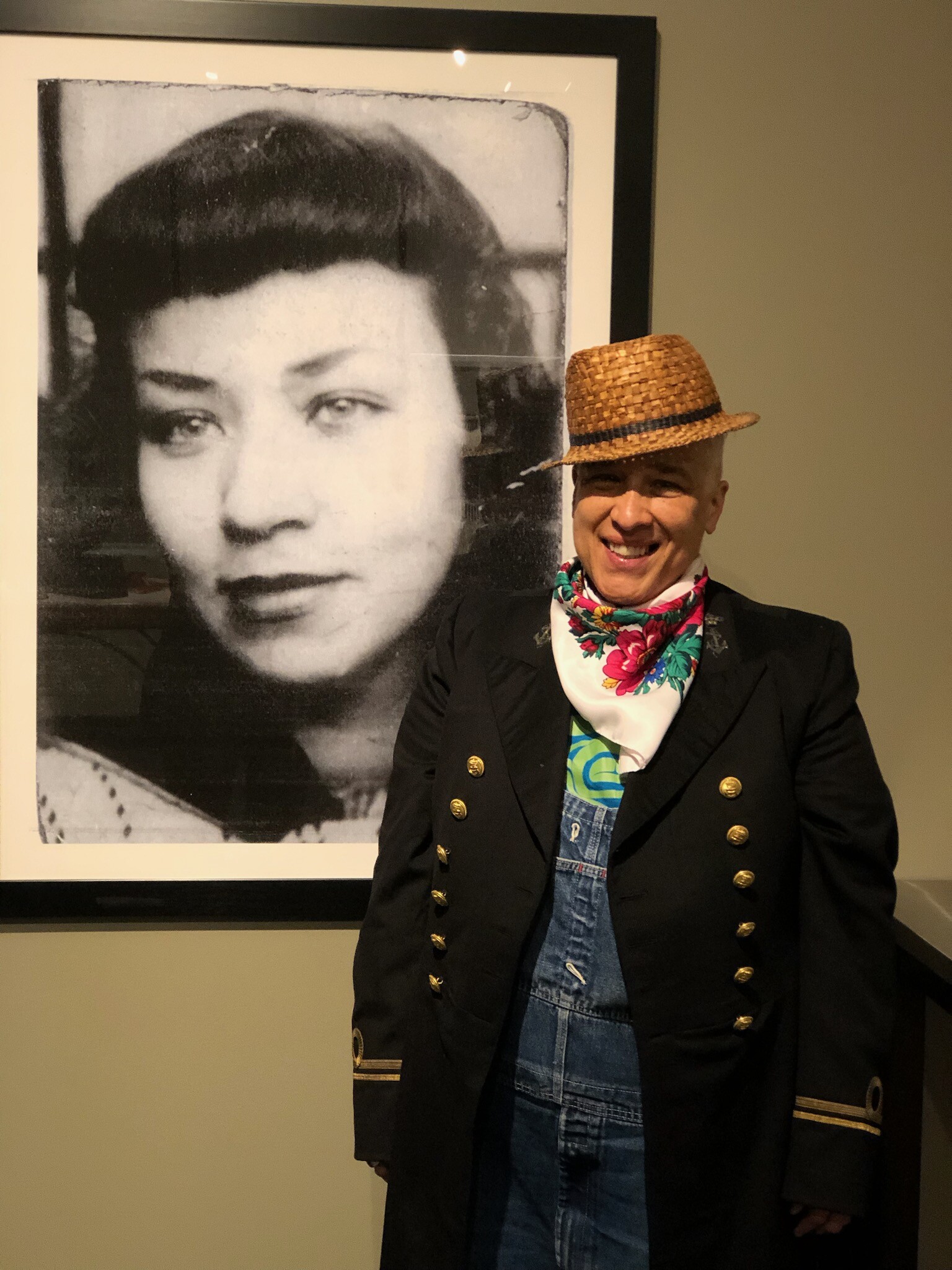 A man in a hat stands next to a portrait of a woman hanging on the wall behind him.