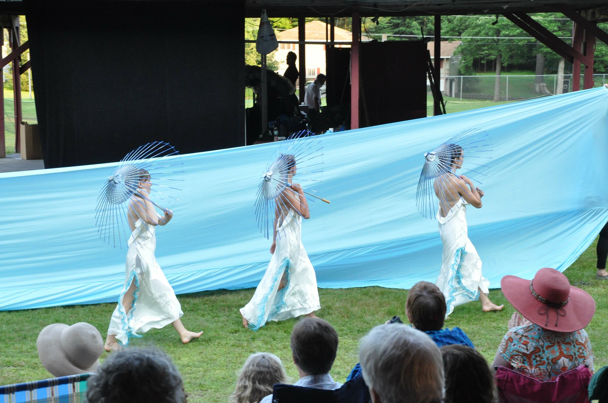 Three performers with mesh parasols walk in front of a large fabric sheet.