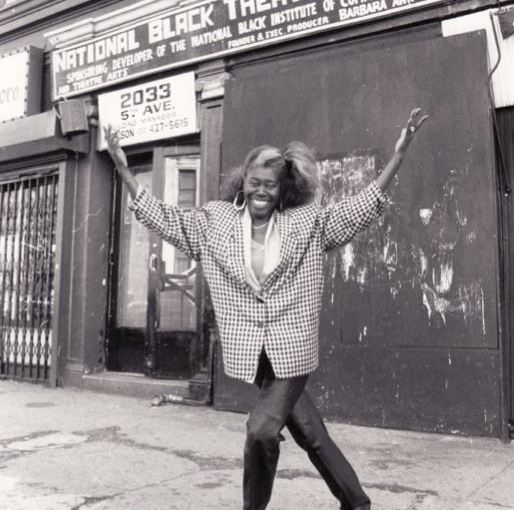 Barbara Teer smiles and gestures to the exterior sign for the National Black Theatre.