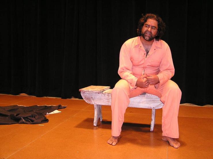 A performer sits on a small bench on stage and appears to speak to the audience.