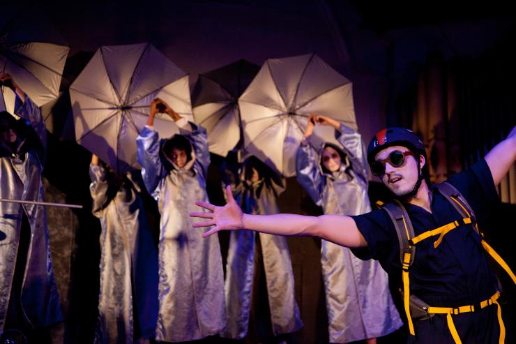 An actor in a helmet and goggles stands with arms outstretched in front of actors with umbrellas on stage.