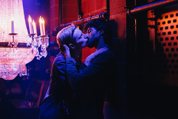 Two men kiss in a dimly lit space onstage.