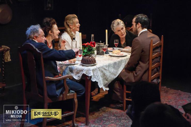 A group of actors sit around a table onstage.
