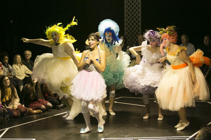 Several actors stand and pose wearing colorful tutus, wigs, and makeup as an audience watches. 