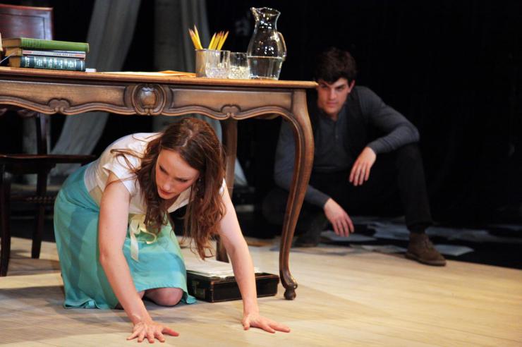 A performer kneels and looks at the stage floor while another performer watches from behind.