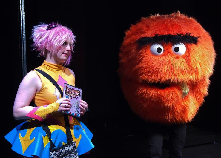 A performer in cosplay stands on-stage with another performer in a large puppet costume.