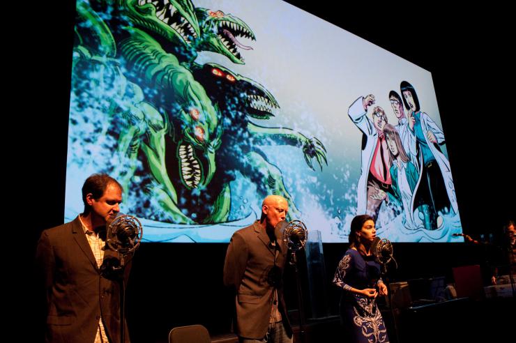 Three performers in front of circular microphones with a comic panel featuring a monster and three scientists behind them.