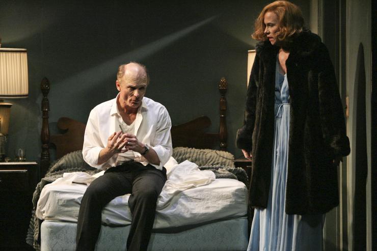 Two actors perform in a play, one sitting on a bed and one standing.