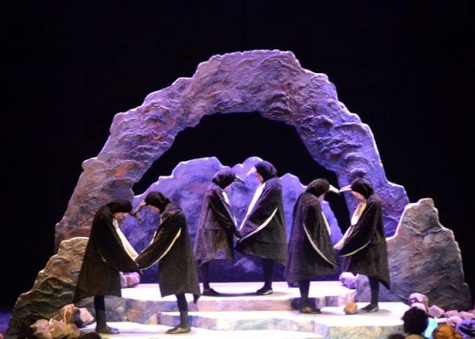 Three couples of actors dressed as penguins holding hands on a set that resembles a rocky outcropping.
