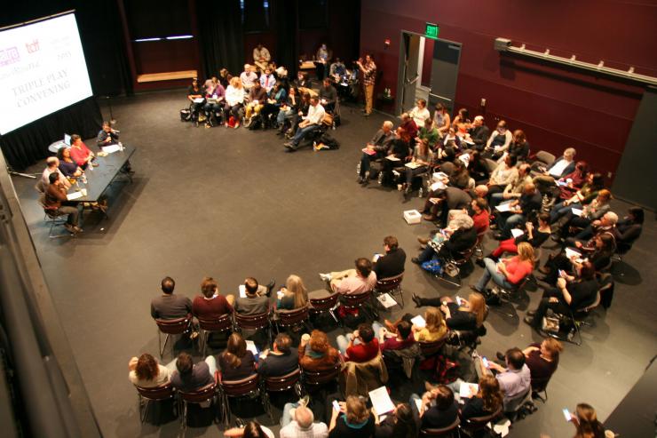 Guests sit in a large semi-circle facing five theatre artists at a table.