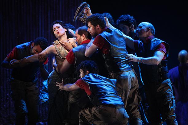 Multiple actors clutch a brightly lit woman onstage.