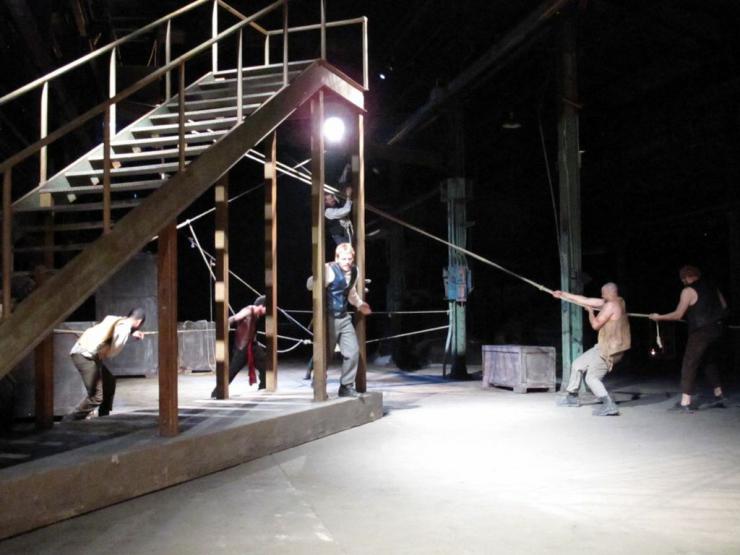 Performers pulling ropes connected to a large wooden staircase on stage. 
