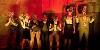 Seven masked performers in semi-formal attire stand in front of a red background.