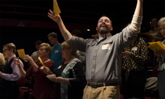 A man looks outward with his arms raise as a choir sings behind him.