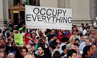 A large group of people with a sign that reads "Occupy Everything" behind them.