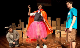 Three people stand on a stage. One kneels on the ground wearing horns, one poses while wearing a pink tutu and orange cape, and other looks out into the audience while wearing a cookie monster t-shirt.
