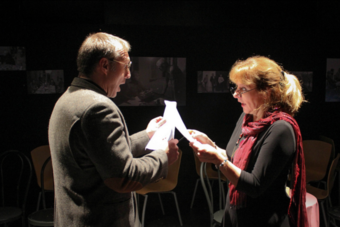 Two actors stand onstage facing each other, reading from sheets of paper.
