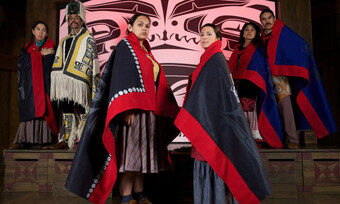 Two native women stand at the center of the photo with their bodies turned toward each other and facing the camera. Their expressions are serious. They are dressed in the cultural wear of their people.