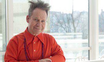 Peter Sellars in a red shirt next to a window.