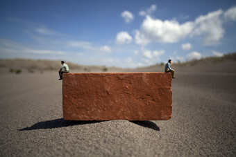 two miniature people balancing on a red brick in a desert.