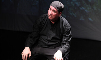 performer sitting, wearing a black button-down and black pants.