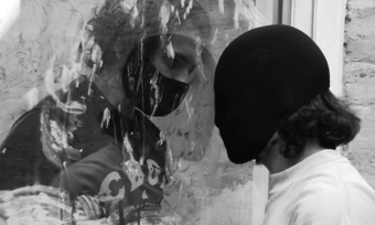 black and white photo of two people in different masks facing each other through glass