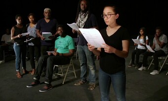 5 actors stand upstage looking at one actor who stands closer to the camera, downstage. They hold scripts and look very somber.