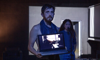 an actor at a microphone stands behind actor holding a computer facing the camera.