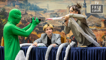 actors in office attire perform with actor in green screen suit onstage.
