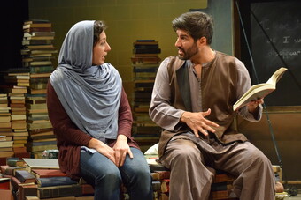 A woman with a blue headscarf, red shirt, and jeans sits next to a man in a brown vest, gray shirt, and tan pants on stacks of books. The man is holding a book open and both people appear to be speaking. They are in front of other piles of books with half of a chalkboard visible in the background.