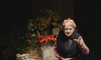 performer portraying an elderly roma woman in front of flowers.