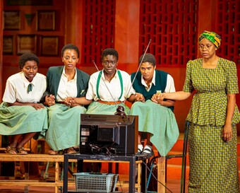 5 Actors on stage looking at a TV Screen. Four actors wear schoolgirl uniforms. One actor stands wearing a green blouse one and skirt 