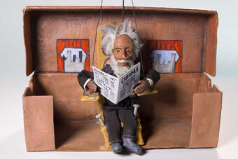 a puppet of frederick douglass sits on a bench reading a newspaper.