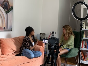 Two women sitting across from each other being filmed in a living room.
