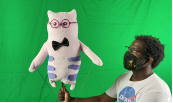 A man holds a puppet above his head in front of a green screen.