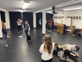 Several actors standing or sitting in a practice space.