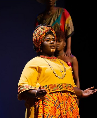 A Black performer wearing patterned headband and yellow shirt on stage