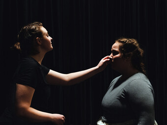 An actor touches the face of another actor while standing on a darkened stage.