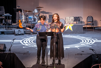 Two women stand on stage in front of music stands on microphones.