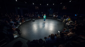 A woman stands and gives a speech on a round stage as the audience watches on.